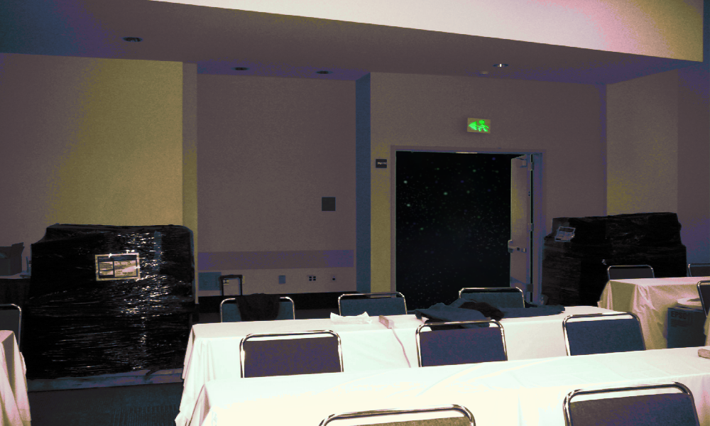 a convention or expo center side room. tables are set up with tableclothes, there is a suitcase in the back, and shipping palettes wrapped in plastic with unknown contents sit in the room. there are at lease two or three rows of tables and three columns. It could  be a panel room. The door leading out of the room opens to an empty tarry and starry black void